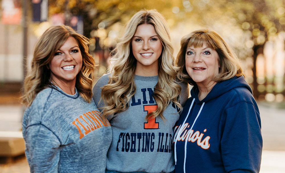 Three generations of women smiling and standing in front of an alumni background