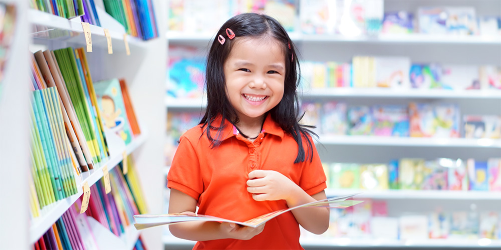 A smiling child with a book in her hand surrounded by other books on shelves