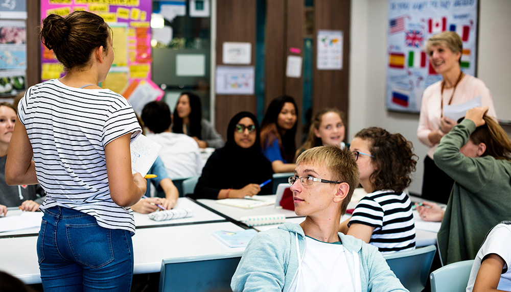 Student standing up giving a presentation to teacher and other students, with various ethnicities involved